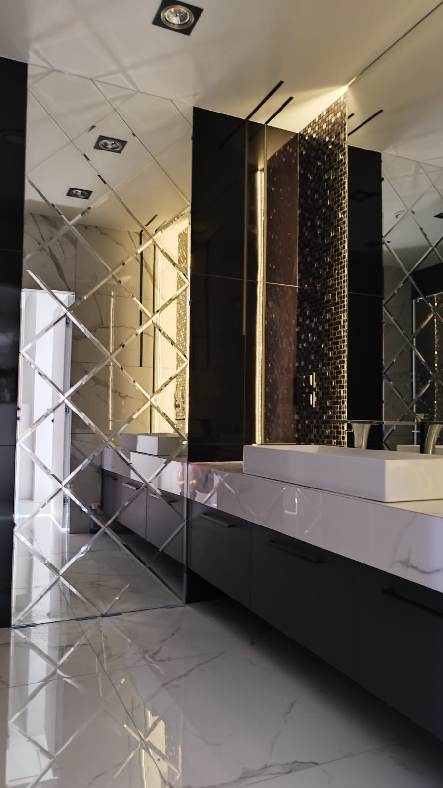 Nowoczesna łazienka z lustrzaną ścianą ułożoną z romboidalnych płytek, białą umywalką nablatową i czarnymi szafkami pod umywalką, na podłodze biały marmur z szarymi żyłami.