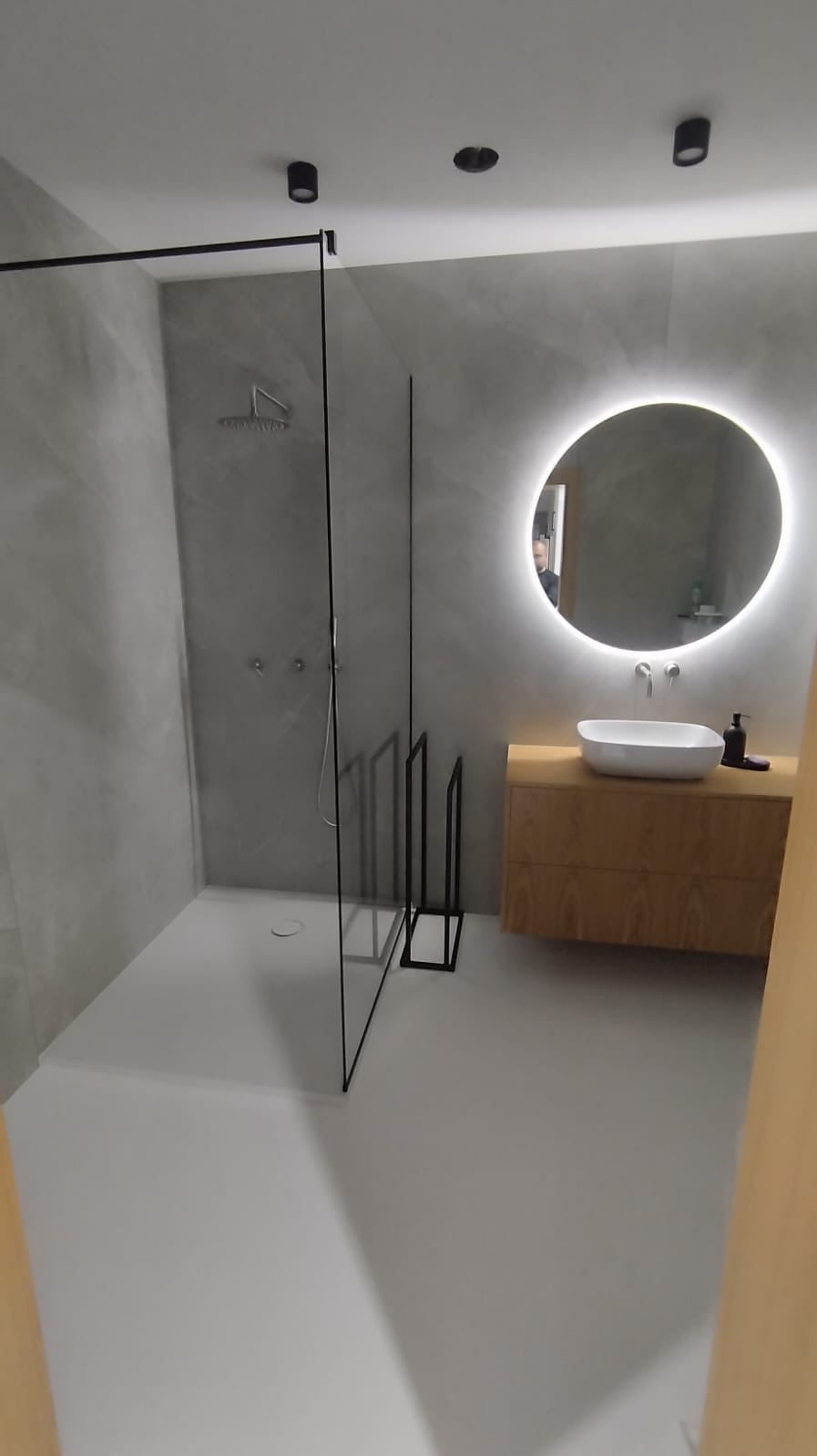 Nowoczesna łazienka z kabiną prysznicową ze szkła hartowanego z czarnymi profilami, okrągłe lustro podświetlane LED, drewniana szafka z umywalką nablatową, szara ściana z efektem betonu.
