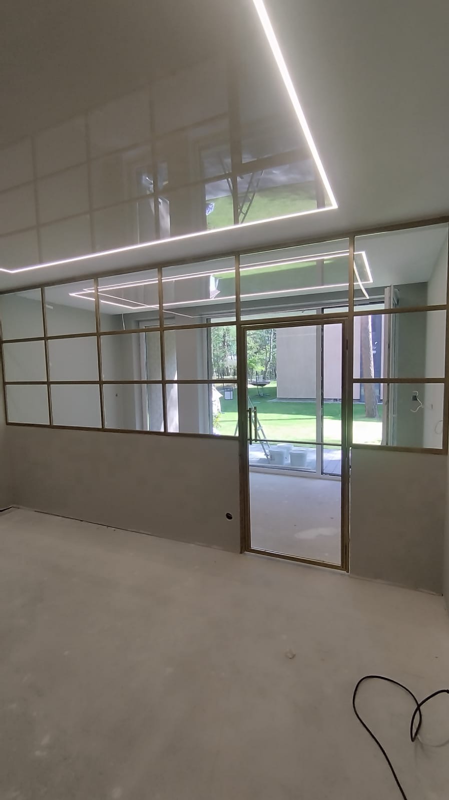 Wnętrze z metalową konstrukcją ścianki działowej ze szklanymi panelami i przesuwnymi drzwiami, widok na ogród przez szklane powierzchnie, sufit z oświetleniem LED.