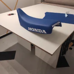 Niebieskie siedzisko motocyklowe marki Honda leży na białym stole roboczym, obok leży kawałek niebieskiego materiału i rolka materiału. Na stole leżą też klucze.