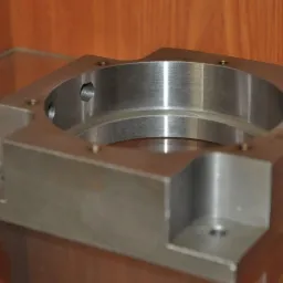 Szara, metalowa obudowa z otworem centralnym i gwintowanymi otworami bocznymi, leżąca na szklanej półce z drewnianym tłem.