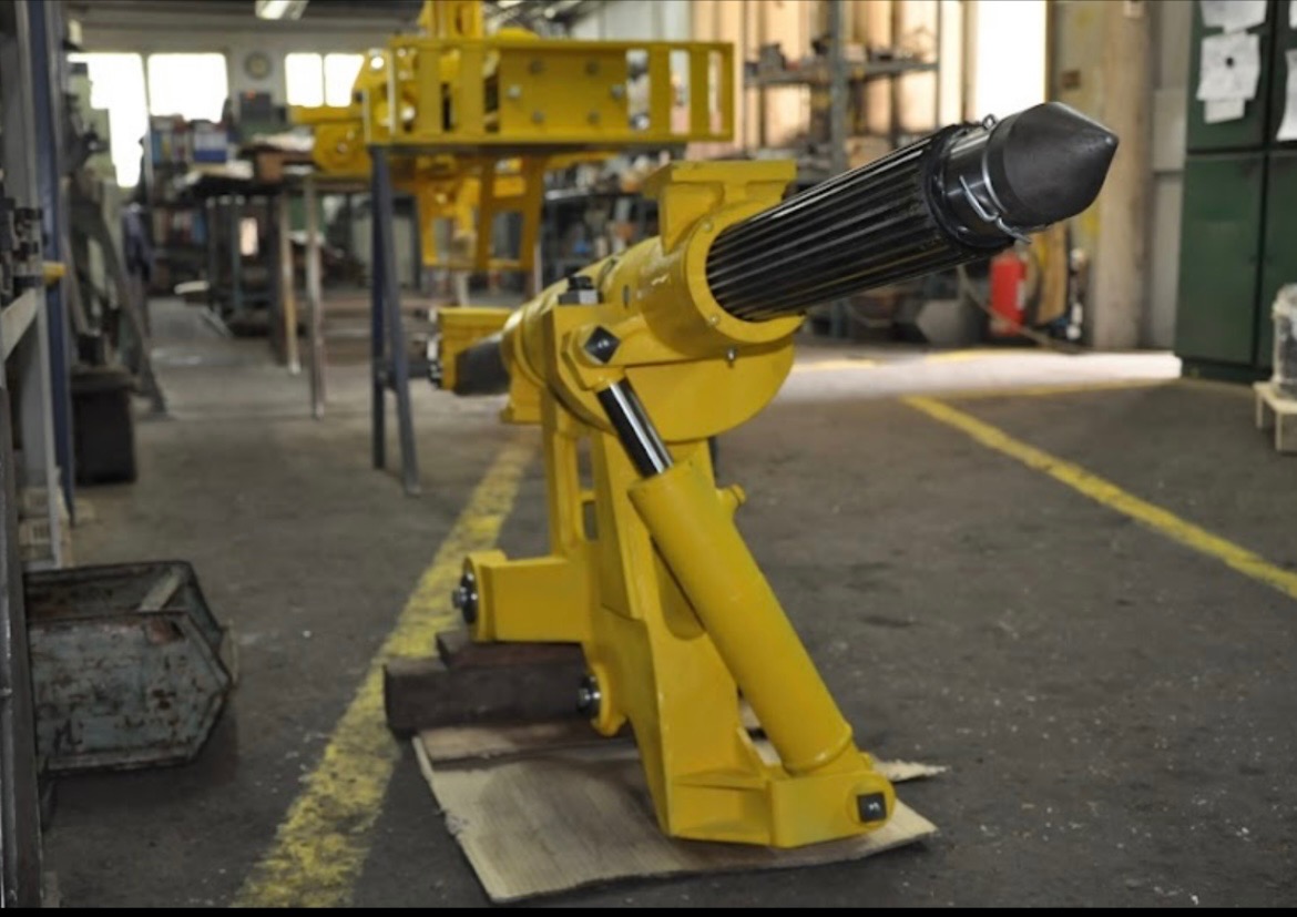 Żółta, hydraulicznie sterowana maszyna z głowicą z prętów, ustawiona na drewnianych deskach w hali produkcyjnej z widocznymi stanowiskami pracy w tle.