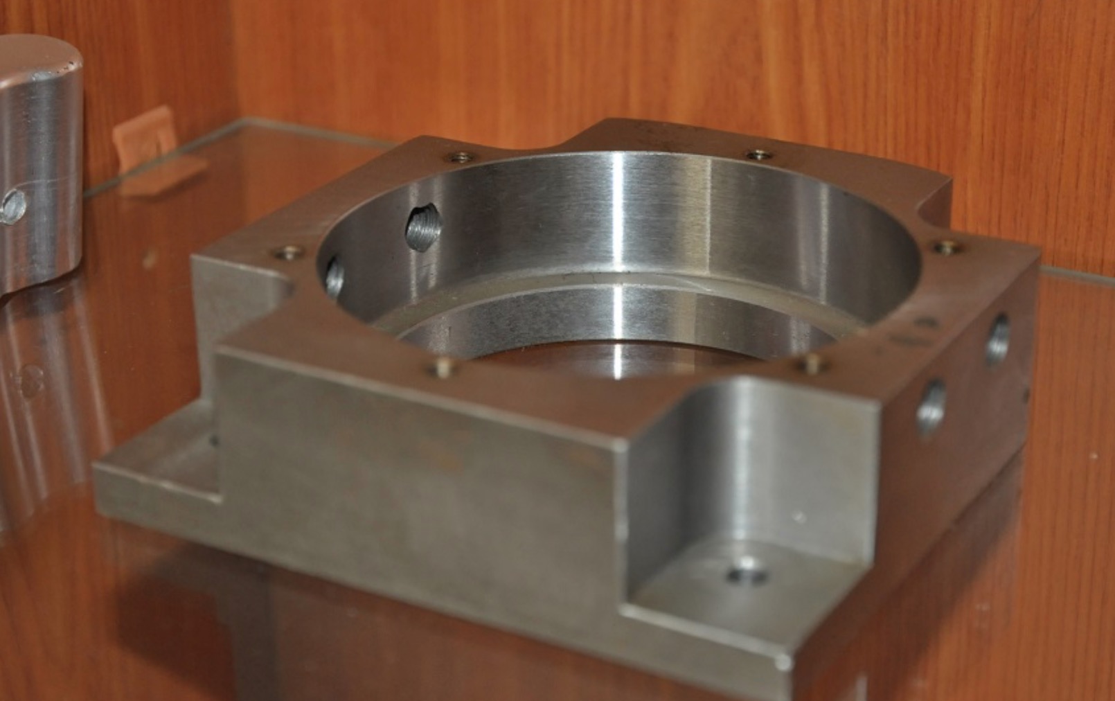 Szara, metalowa obudowa z otworem centralnym i gwintowanymi otworami bocznymi, leżąca na szklanej półce z drewnianym tłem.