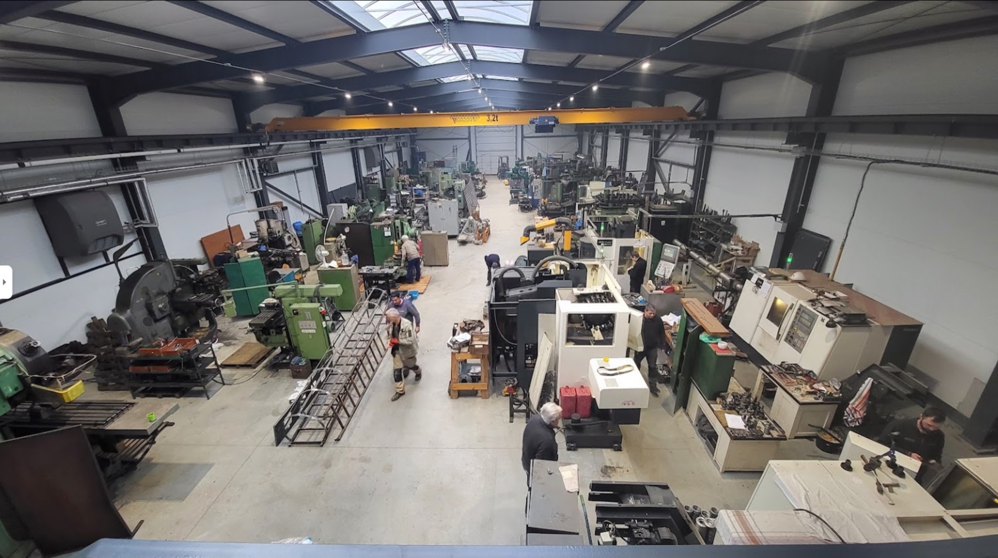 Szeroki kadr hali produkcyjnej z licznymi maszynami do obróbki metali, w tym tokarkami i frezarkami CNC, widoczne suwnice pod stropem, pracownicy w trakcie pracy.