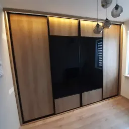 Duża szafa przesuwna z drewnianymi panelami i czarnymi lustrzanymi drzwiami, oświetlona od góry, wiszące szklane lampy, kalendarz na ścianie, jasna podłoga i okno z żaluzjami.