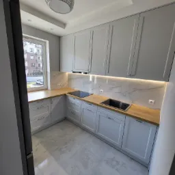 Jasnoszara kuchnia z drewnianym blatem, oświetleniem podszafkowym LED i okrągłą lampą sufitową w tle okna.