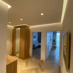 Nowoczesny przedpokój z drewnianą podłogą w jodełkę, szafą z lustrem i podświetlanym sufitem, widok na salon i pokój.