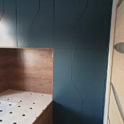 Ciemnogranatowe szafy z geometrycznymi wgłębieniami na frontach, połączone z drewnianą konstrukcją antresoli z widocznymi otworami na materac, wkomponowane w jasne wnętrze.