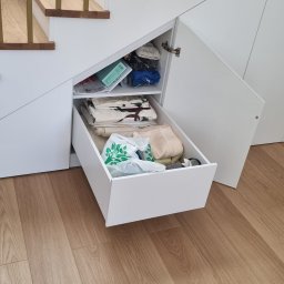 SONO MEBLE - Biała szafka pod schodami z otwartą szufladą i półką, wypełniona tekstyliami i torbami. Widoczne drewniane schody i panele podłogowe.