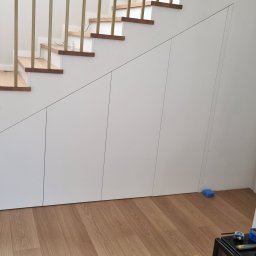 SONO MEBLE - Biała szafa wnękowa pod schodami z drewnianymi stopniami i złotymi barierkami. Widoczna podłoga z paneli i narzędzia na szafce w rogu.