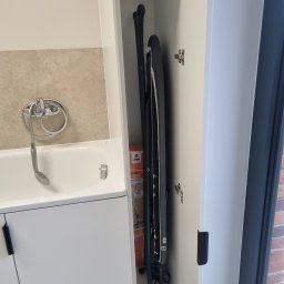 SONO MEBLE - Biała szafa wnękowa z deską do prasowania w łazience, obok wanny z szarą mozaiką na ścianie i oknem. Ukryte przechowywanie w nowoczesnym stylu.