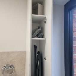 SONO MEBLE - Wnętrze białej szafy wnękowej z półkami, na których znajdują się tekstylny pojemnik, deska do prasowania i suszarka. Widoczna ściana z beżowymi płytkami i bateria.