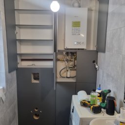 SONO MEBLE - Szara zabudowa w łazience z otwartymi szafkami, widoczne rury i narzędzia na pralce. Montaż szafki z kotłem w łazience w trakcie pracy.