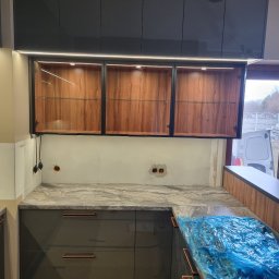 SONO MEBLE - Częściowo zamontowana kuchnia na wymiar: szare szafki dolne z marmurowym blatem, górne przeszklone szafki z oświetleniem LED. Widoczne niedokończone ściany.