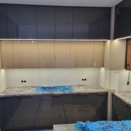 SONO MEBLE - Nowoczesna kuchnia z szarymi szafkami i blatem imitującym marmur, oświetlona listwami LED. Blat częściowo zakryty folią ochronną. Widoczne gniazdka elektryczne w ścianie.