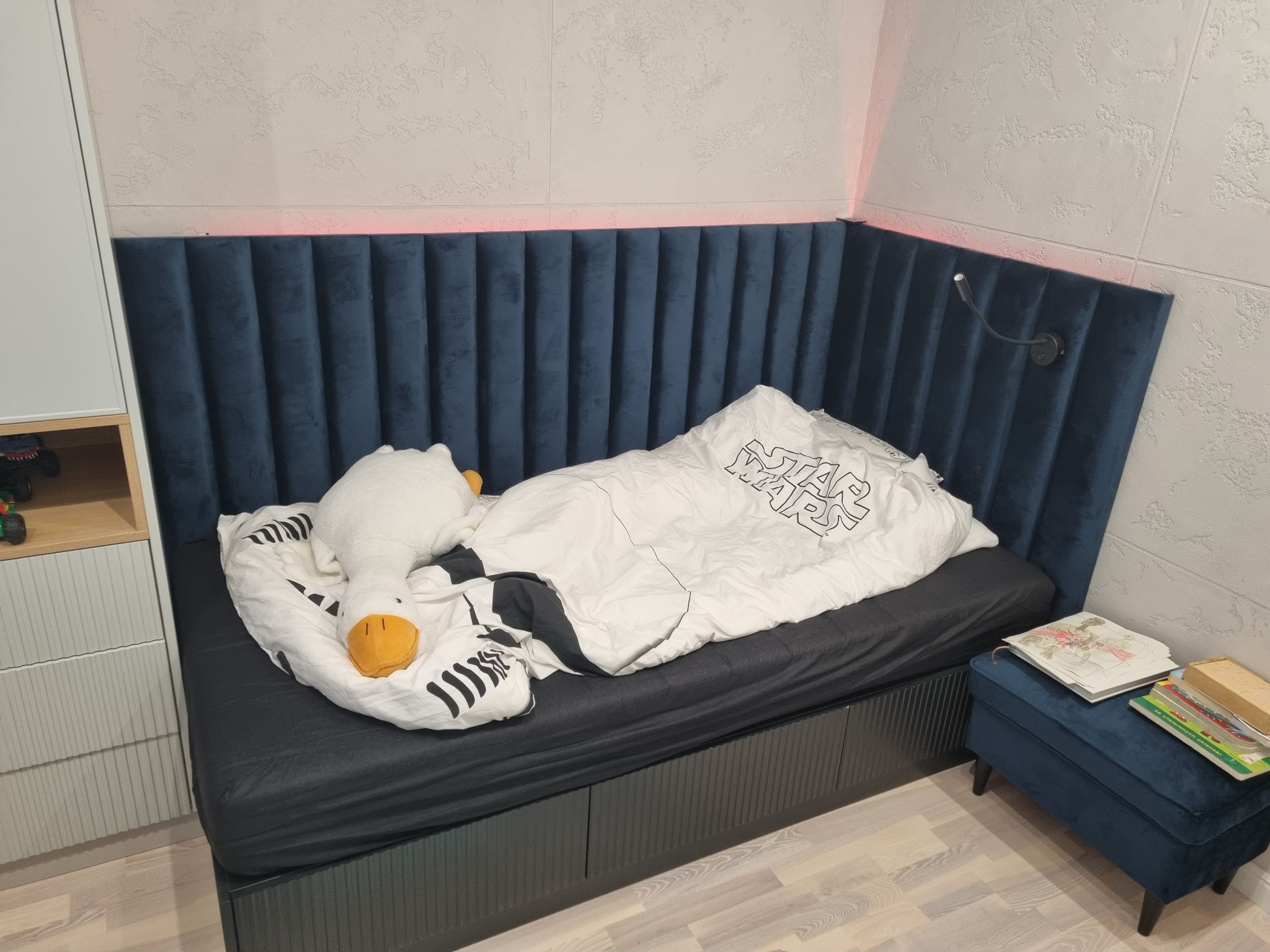 Niebieskie, tapicerowane wezgłowie łóżka z podświetleniem LED, w pokoju dziecięcym. Obok łóżka szafka nocna z książkami. Na łóżku pościel z motywem Star Wars i pluszowa kaczka.