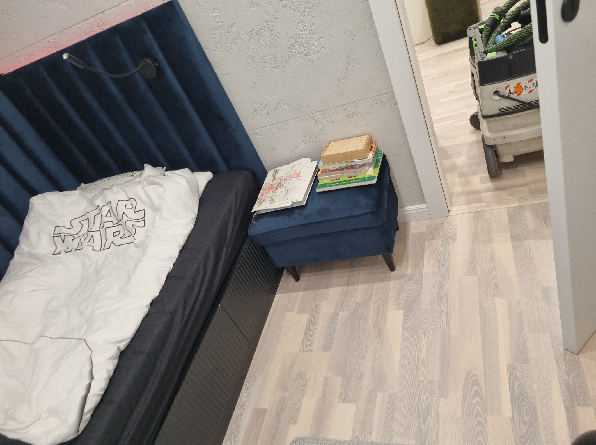 Niebieskie, tapicerowane wezgłowie łóżka z lampką, obok pufa z książkami. W tle odkurzacz przemysłowy i podłoga z paneli w jasnym odcieniu drewna.