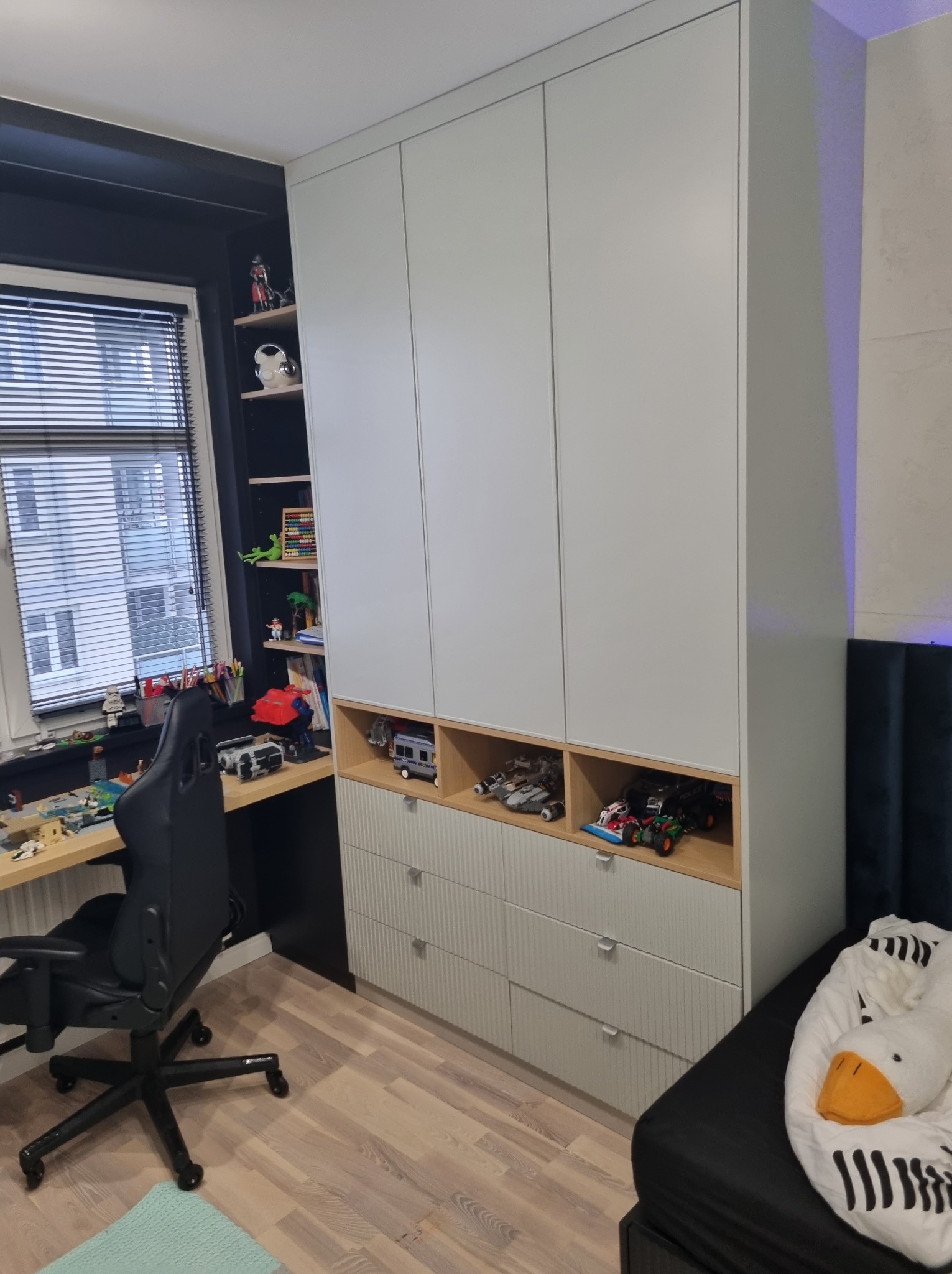 Jasna szafa z szufladami i otwartymi półkami, wypełnionymi zabawkami, w pokoju dziecięcym. Szafa zintegrowana z biurkiem, krzesło obrotowe.