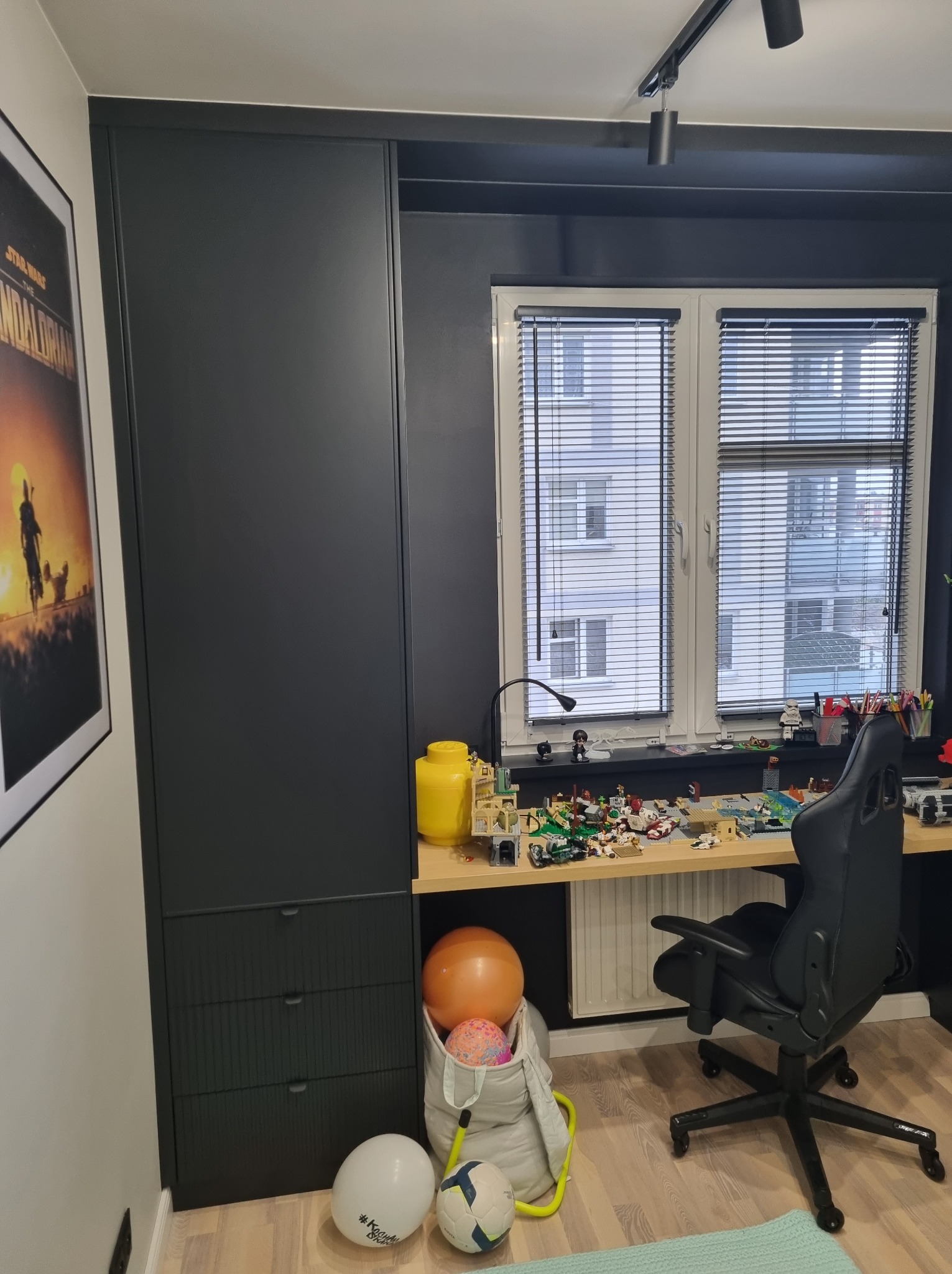 Nowoczesny pokój dziecięcy z czarną szafą z ryflowanymi frontami, biurkiem z zabawkami Lego i oknem z żaluzjami. Minimalistyczny design z akcentami kolorystycznymi.