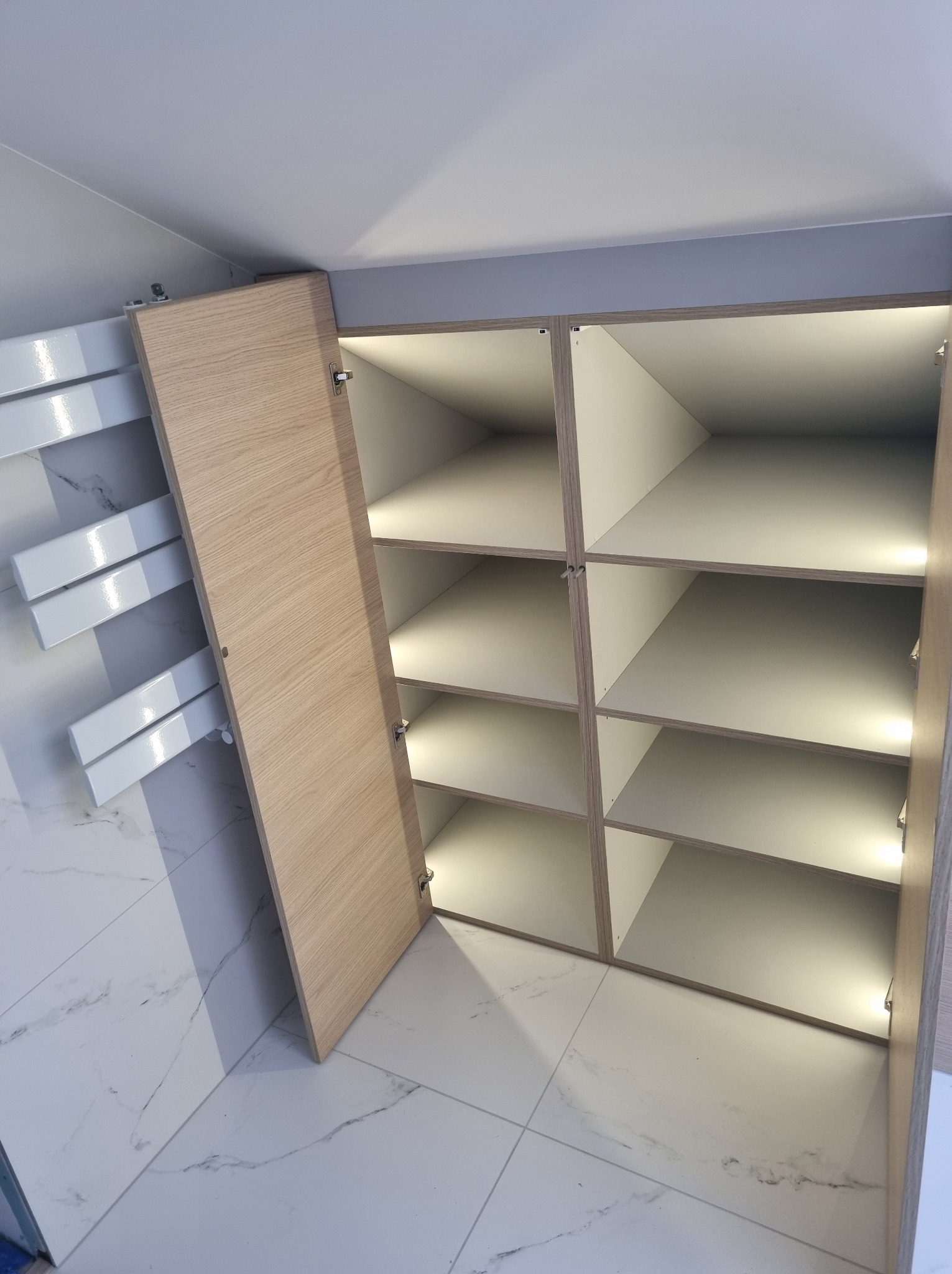 Otwarta szafa wnękowa z oświetleniem LED, jasne drewno i białe wnętrze, widok z dołu, marmurowe płytki podłogowe.