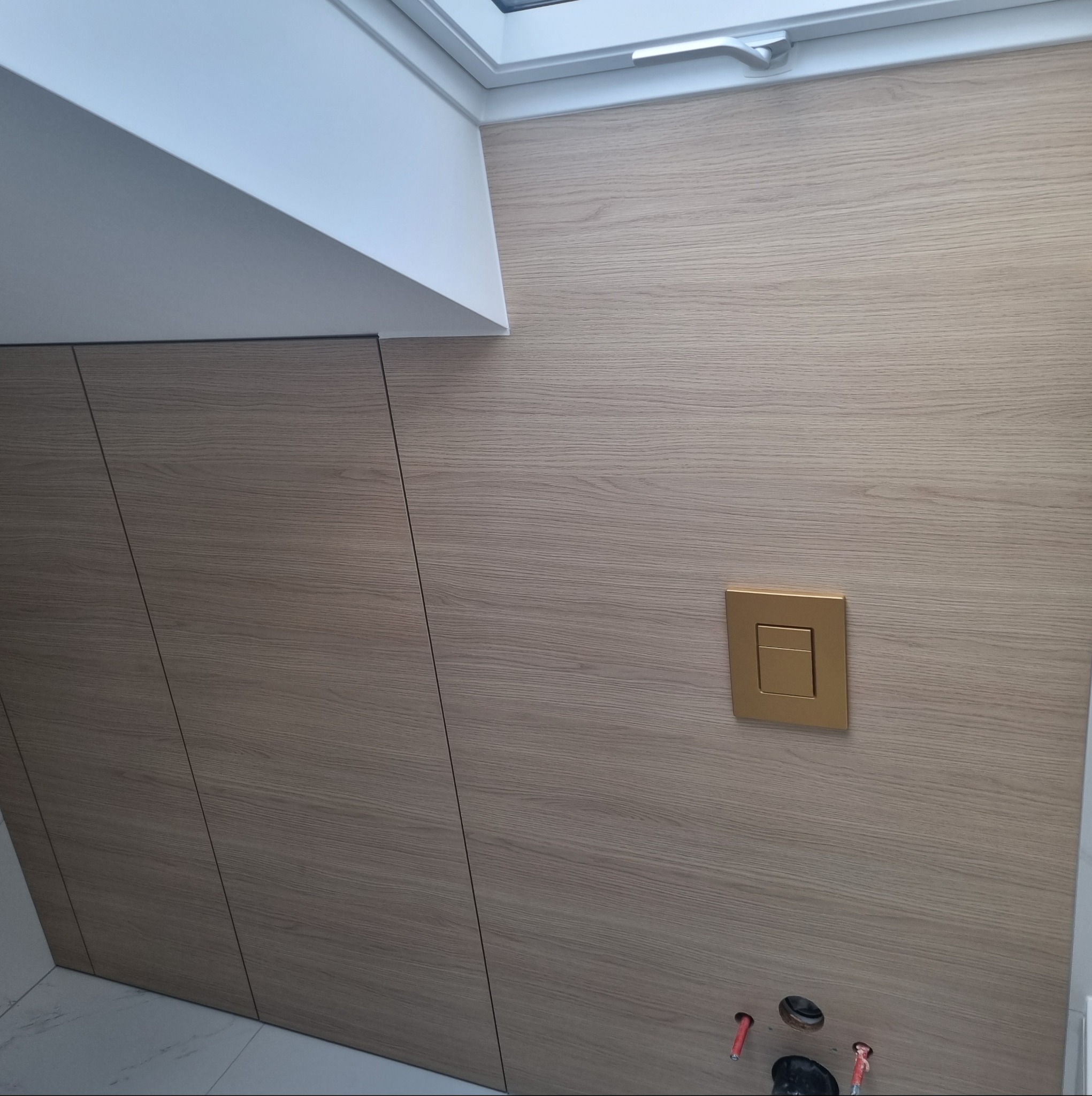 Drewnopodobne panele ścienne z ukrytymi drzwiami i złotym przyciskiem spłuczki pod oknem dachowym. Minimalistyczne wykończenie łazienki.