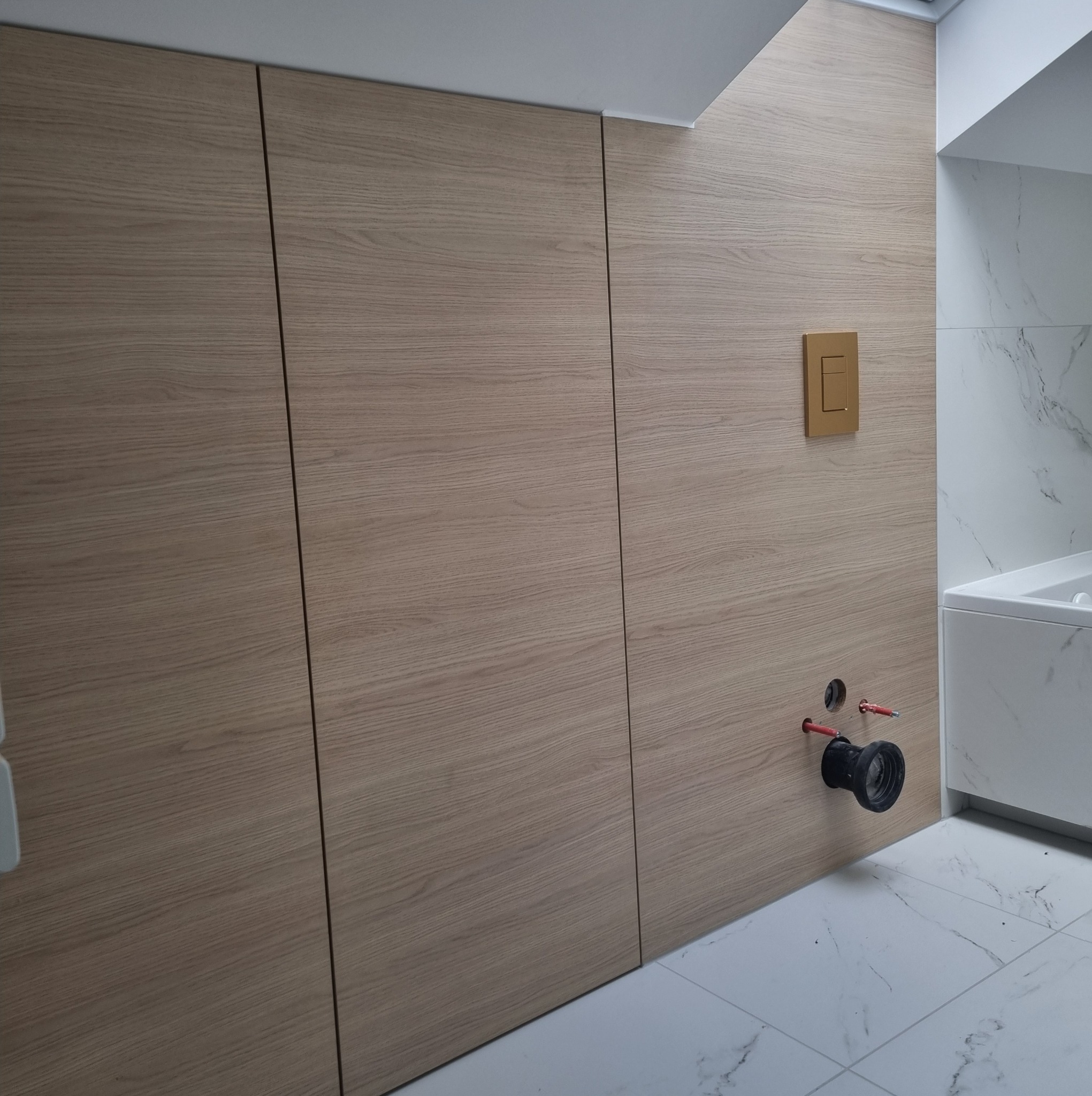 Bordowa szafa wnękowa z drewnianym wykończeniem, montaż odpływu kanalizacyjnego w łazience, widoczny przycisk spłuczki w kolorze złotym.