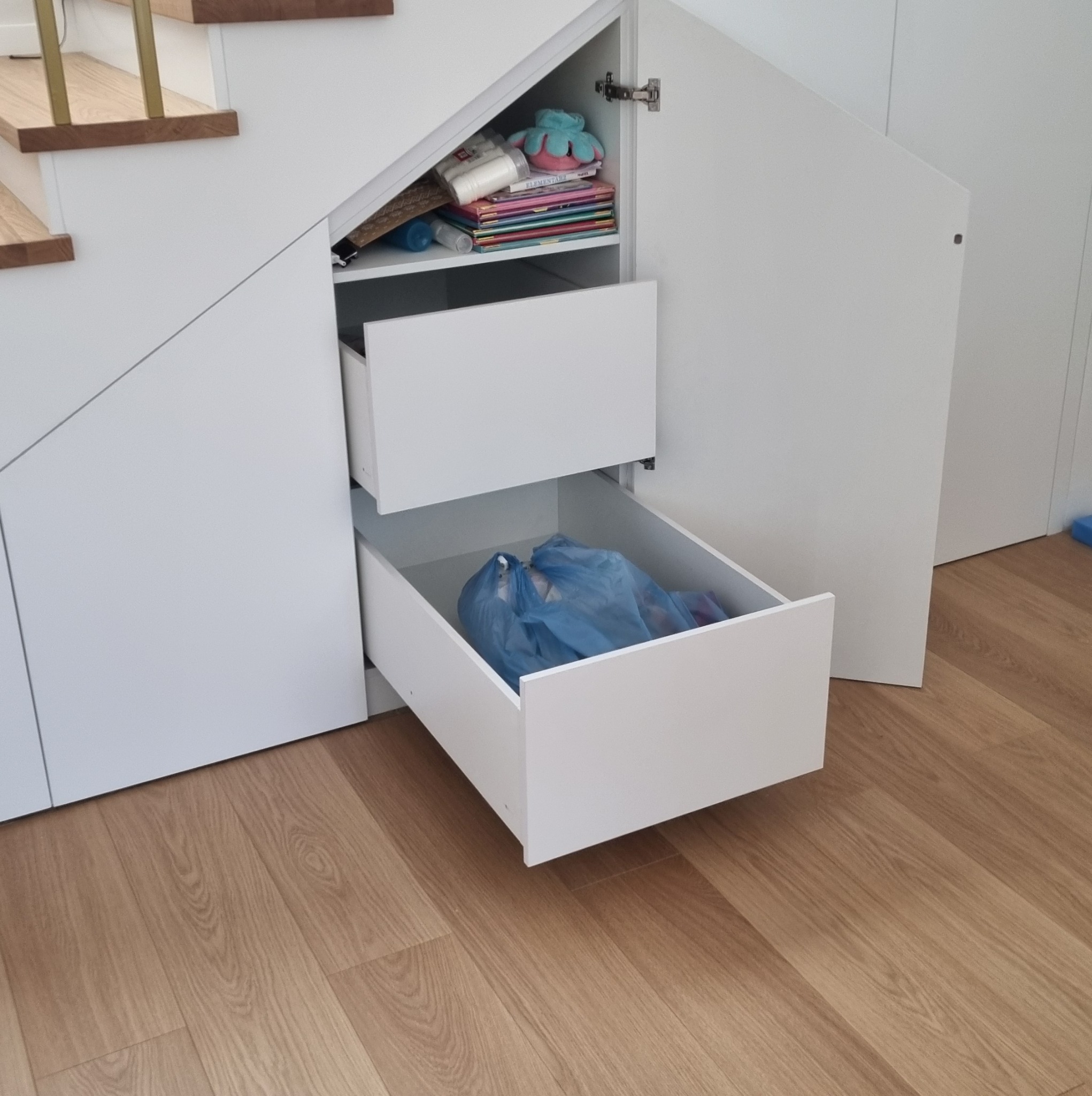 Biała zabudowa pod schodami z otwartymi szufladami i półkami. Widoczne przedmioty wewnątrz. Jasna drewniana podłoga. Ujęcie z dołu.
