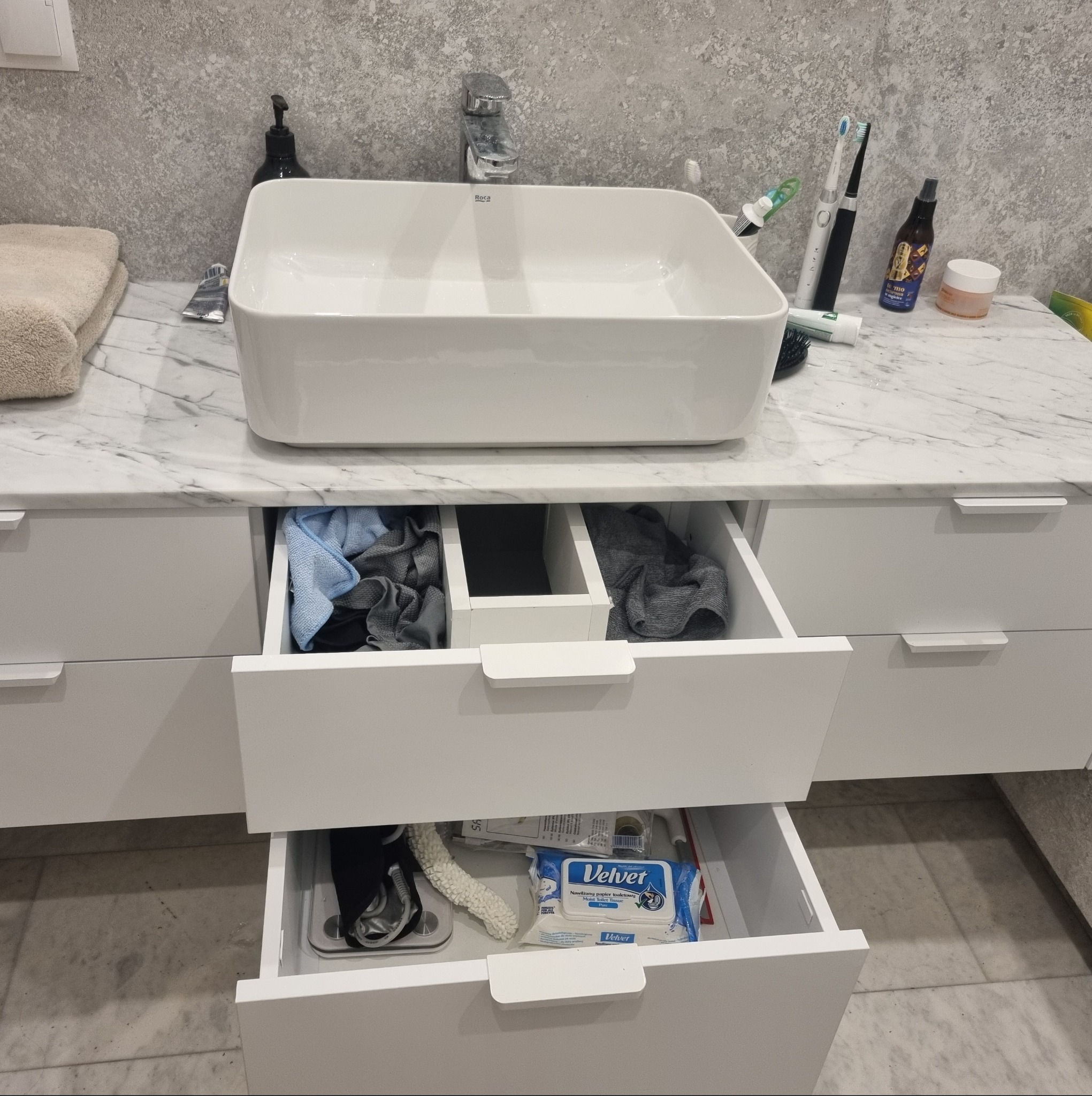 Biała, prostokątna umywalka nablatowa Roca na blacie z marmuru. Otwarta szuflada z tekstyliami i akcesoriami łazienkowymi. Minimalistyczny design.