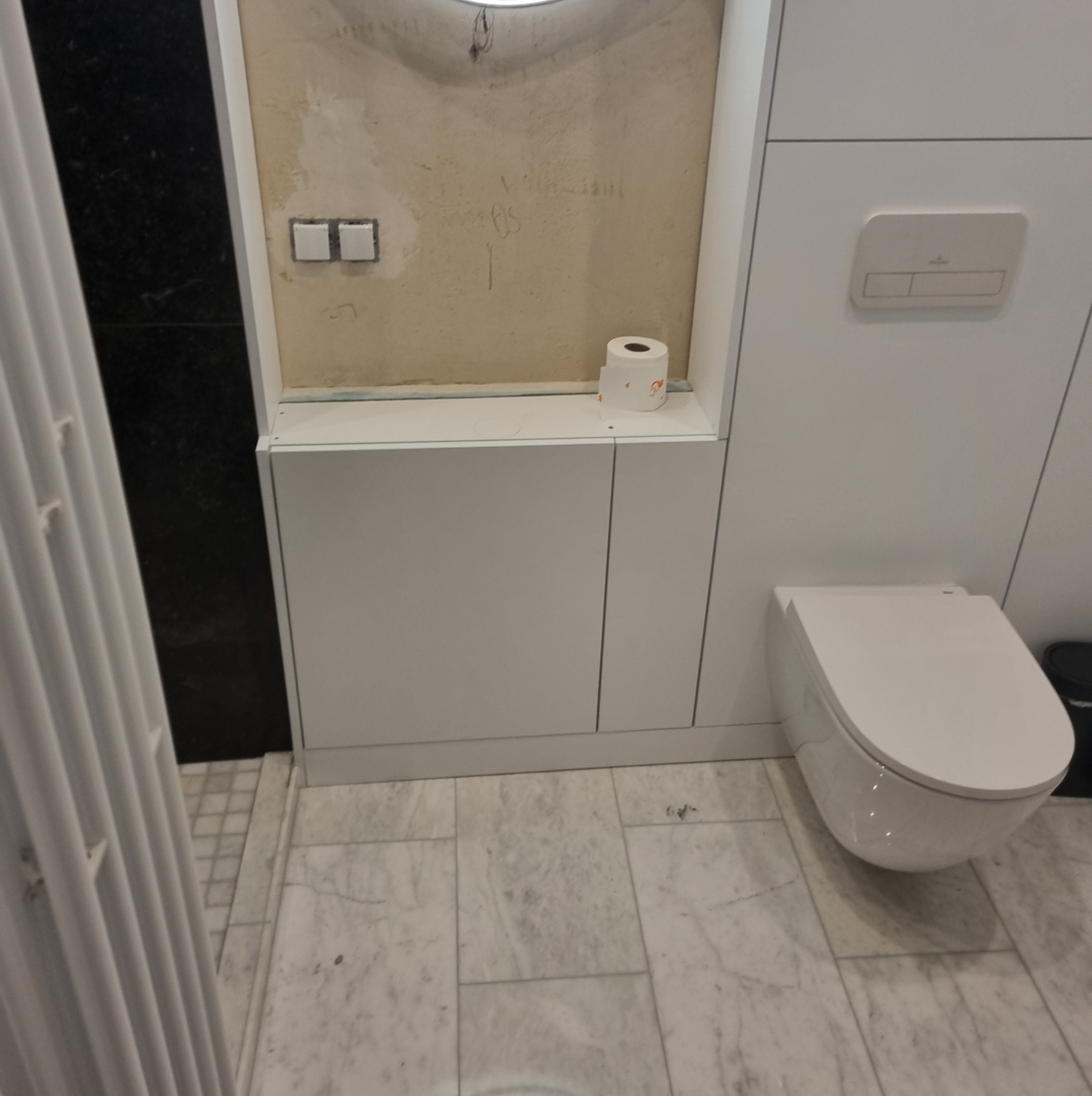 Biała zabudowa WC z szafkami i półką, wisząca toaleta. Ściana w trakcie wykończenia z widocznymi instalacjami. Marmurowe płytki podłogowe.