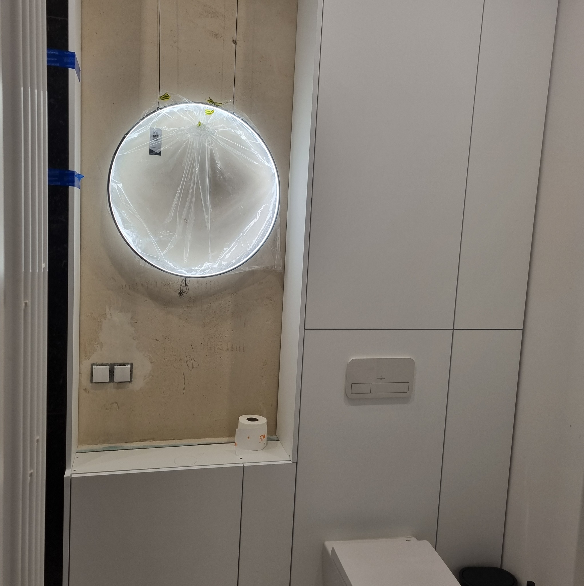 Biała zabudowa łazienkowa z okrągłym lustrem LED w folii ochronnej. Widoczne surowe ściany i instalacja elektryczna. Minimalistyczny design w trakcie montażu.