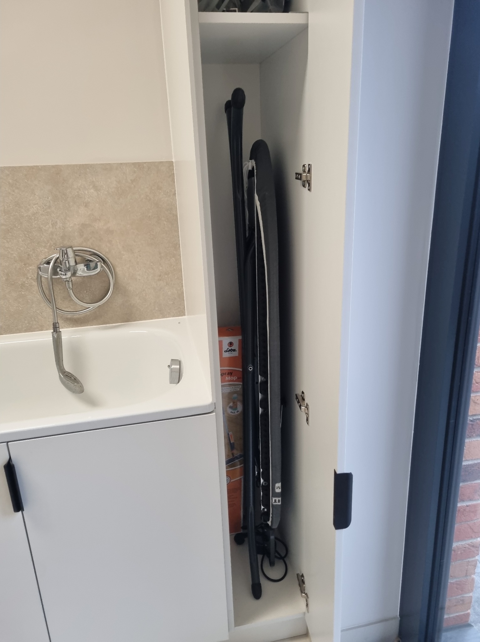 Biała szafa wnękowa z deską do prasowania w łazience, obok wanny z szarą mozaiką na ścianie i oknem. Ukryte przechowywanie w nowoczesnym stylu.