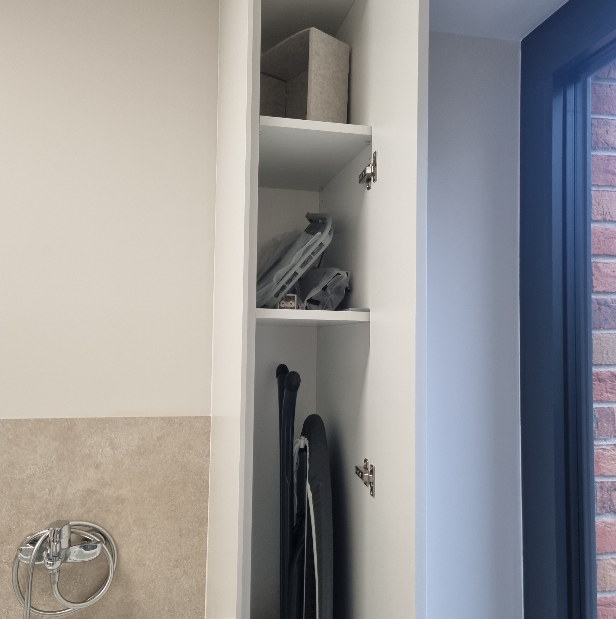 Wnętrze białej szafy wnękowej z półkami, na których znajdują się tekstylny pojemnik, deska do prasowania i suszarka. Widoczna ściana z beżowymi płytkami i bateria.