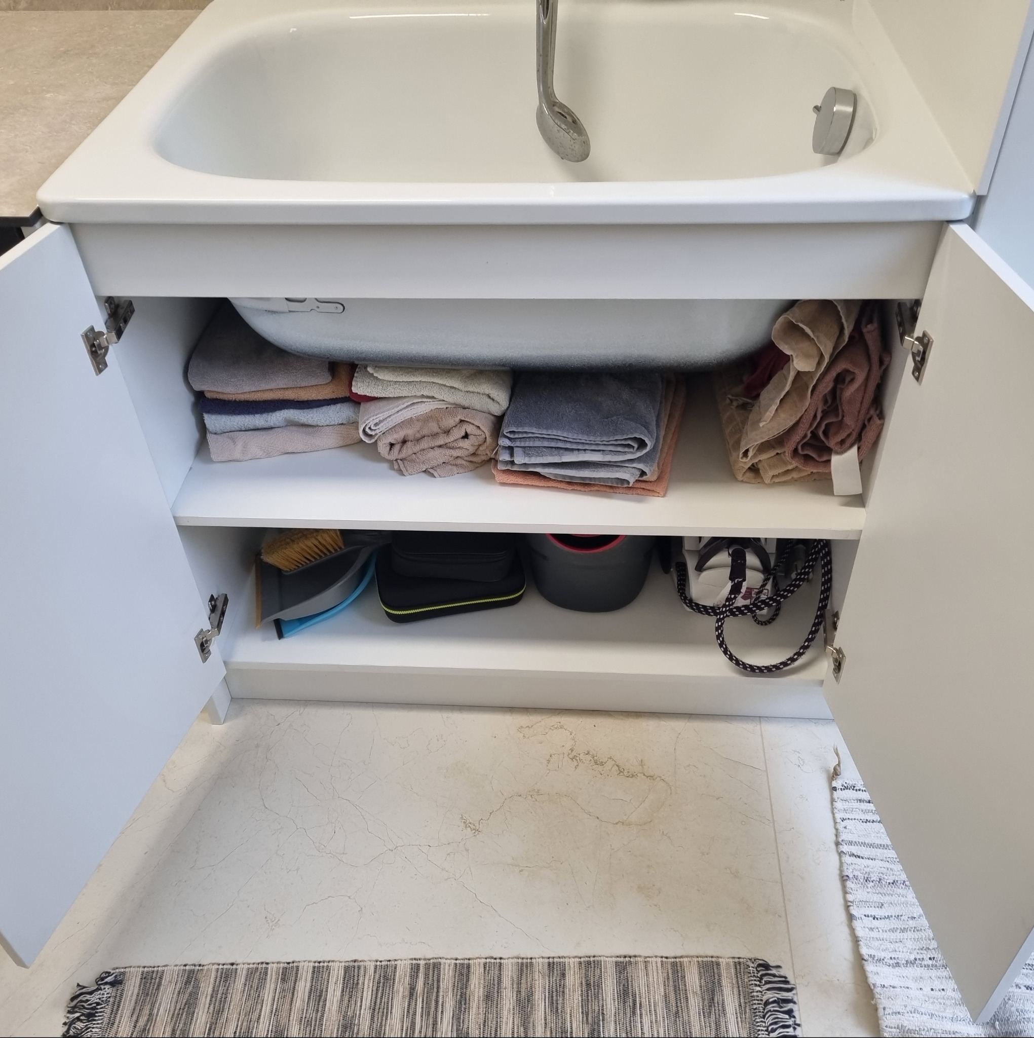 Biała szafka pod umywalką z otwartymi drzwiczkami, widoczne półki z ręcznikami, szczotką, żelazkiem i innymi akcesoriami. Marmurowa podłoga i dywanik.