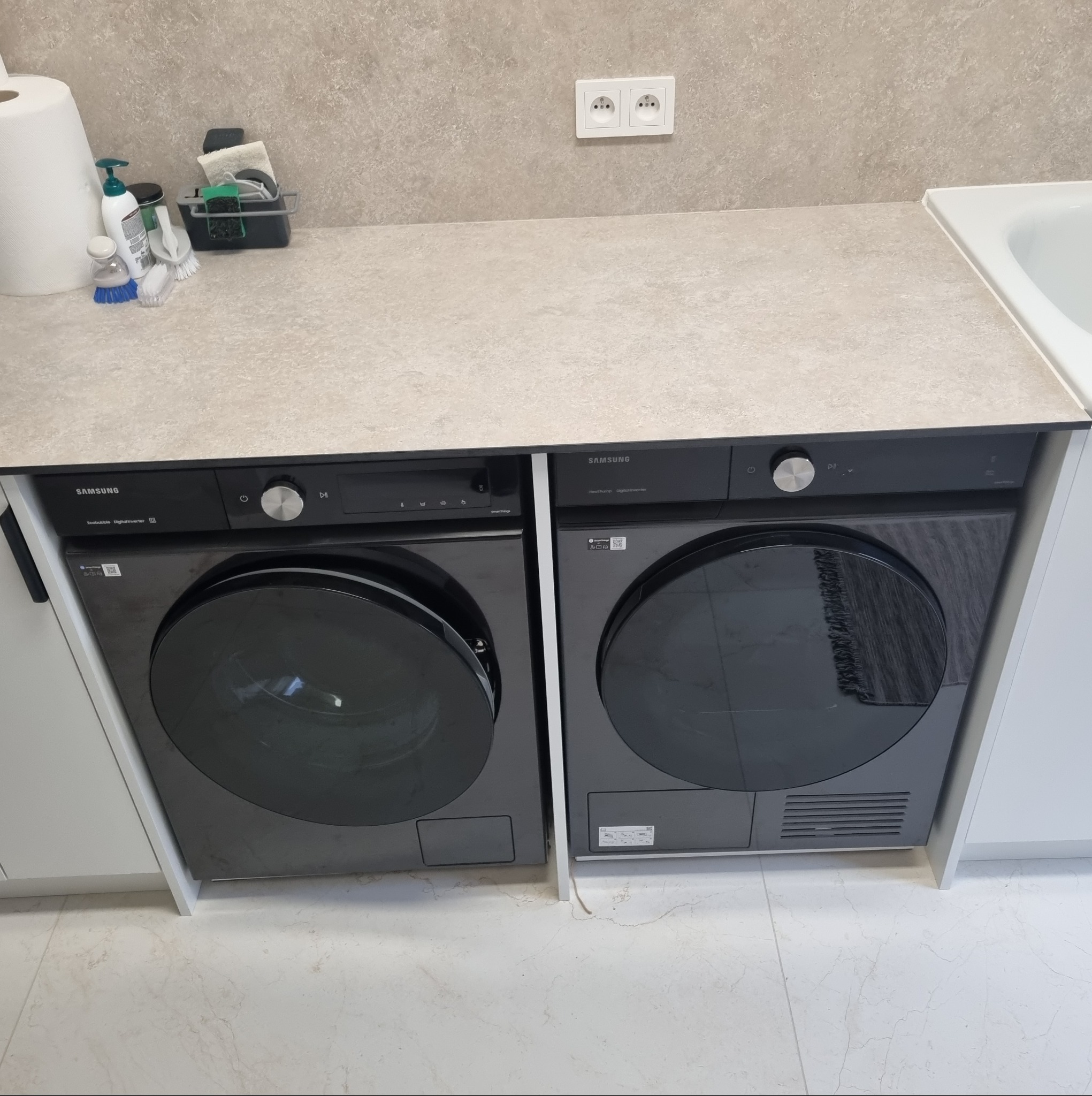 Pralka i suszarka zabudowane pod blatem w łazience w Mazowieckim. Sprzęt marki Samsung, fronty szafek białe, blat w odcieniach beżu. Widoczne gniazdko elektryczne.