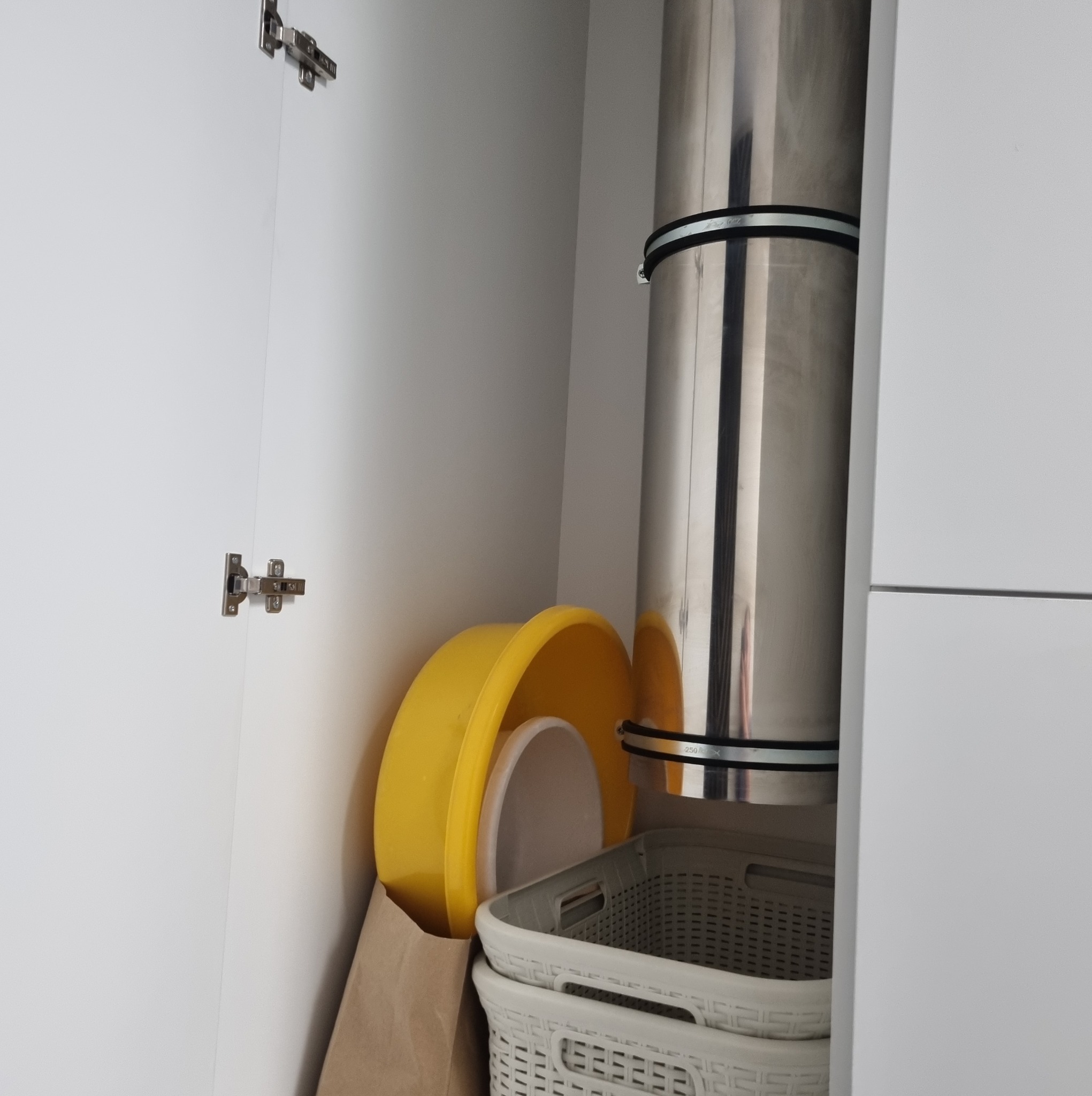Wnętrze białej szafki z metalowym kominem okapu, żółtą miską, papierową torbą i koszykami. Widoczne zawiasy drzwiczek.
