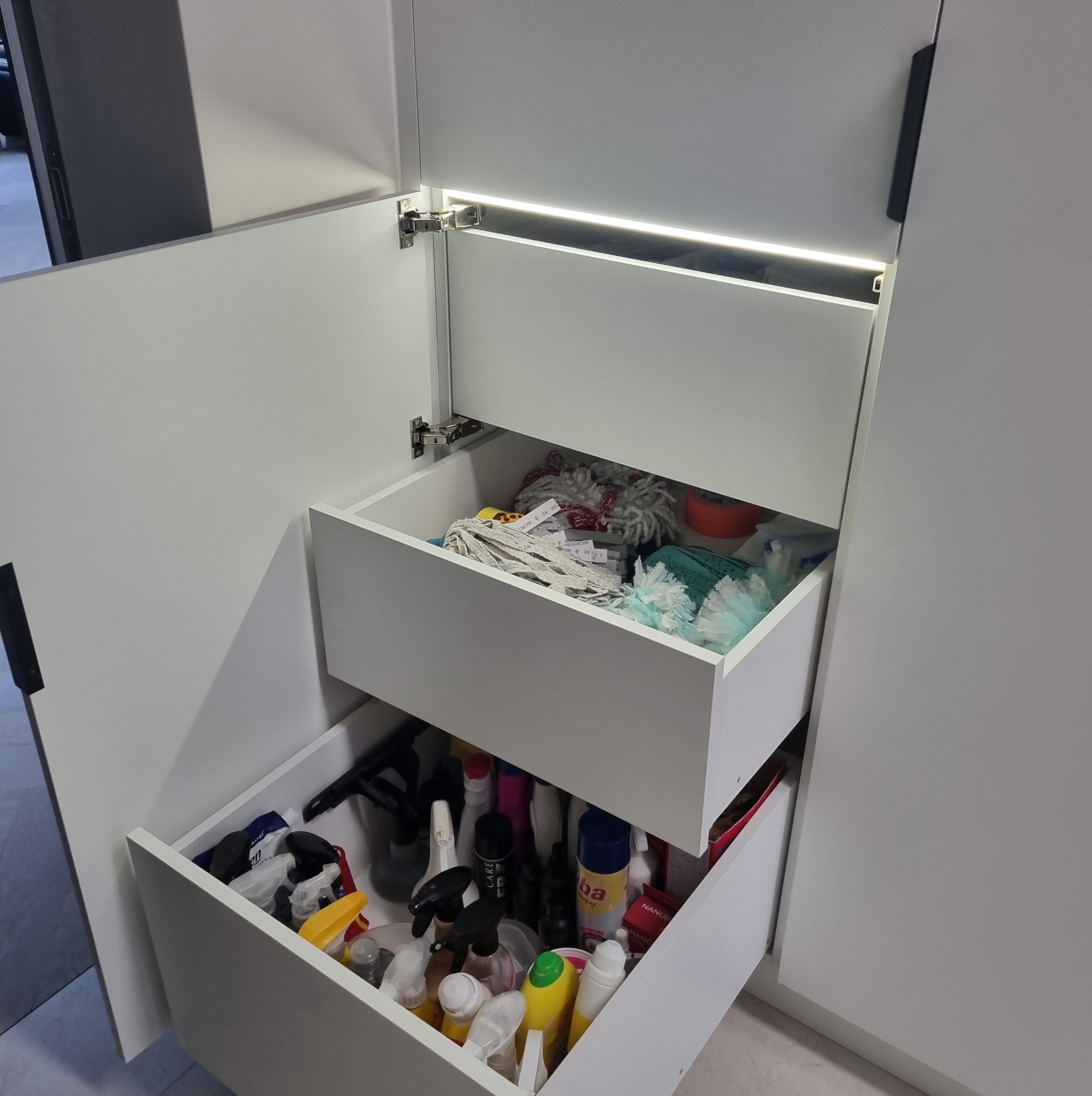 Otwarta, biała szafka z szufladami, oświetlona od wewnątrz, wypełniona środkami czystości i akcesoriami do sprzątania. Widoczne zawiasy i uchwyty.