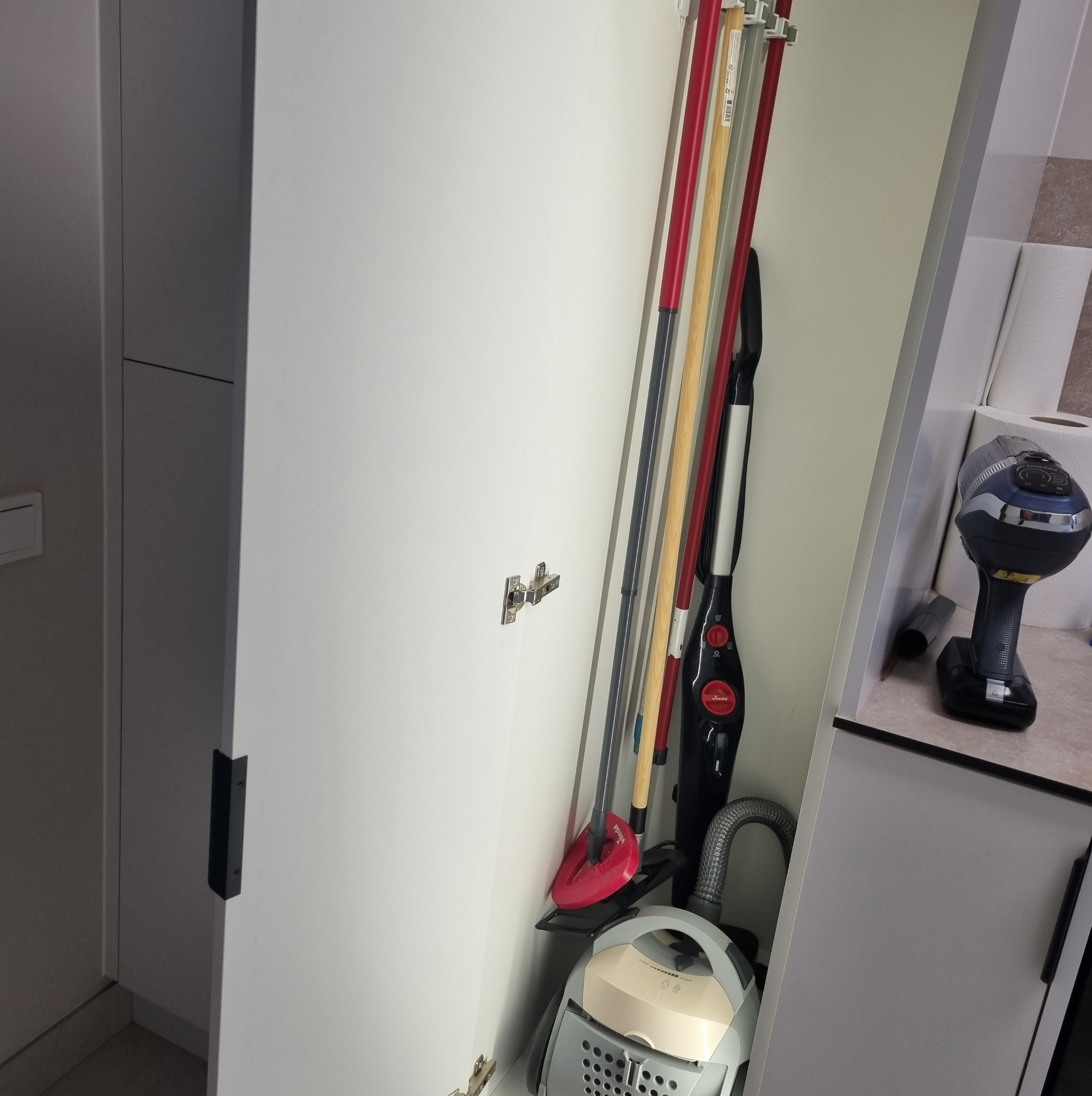 Wnętrze białej szafy na wymiar z różnymi sprzętami do sprzątania: odkurzacz, mop, szczotki. Widoczne zawiasy i uchwyt szafki. Ujęcie z bliska, neutralne oświetlenie.