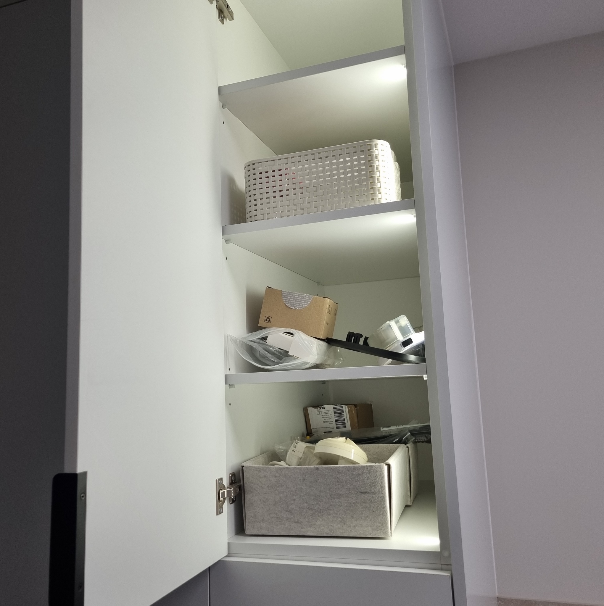 Otwarta, biała szafka z półkami podświetlonymi LED, wypełniona pojemnikami i akcesoriami. Widoczne zawiasy i czarny uchwyt. Minimalistyczny design.