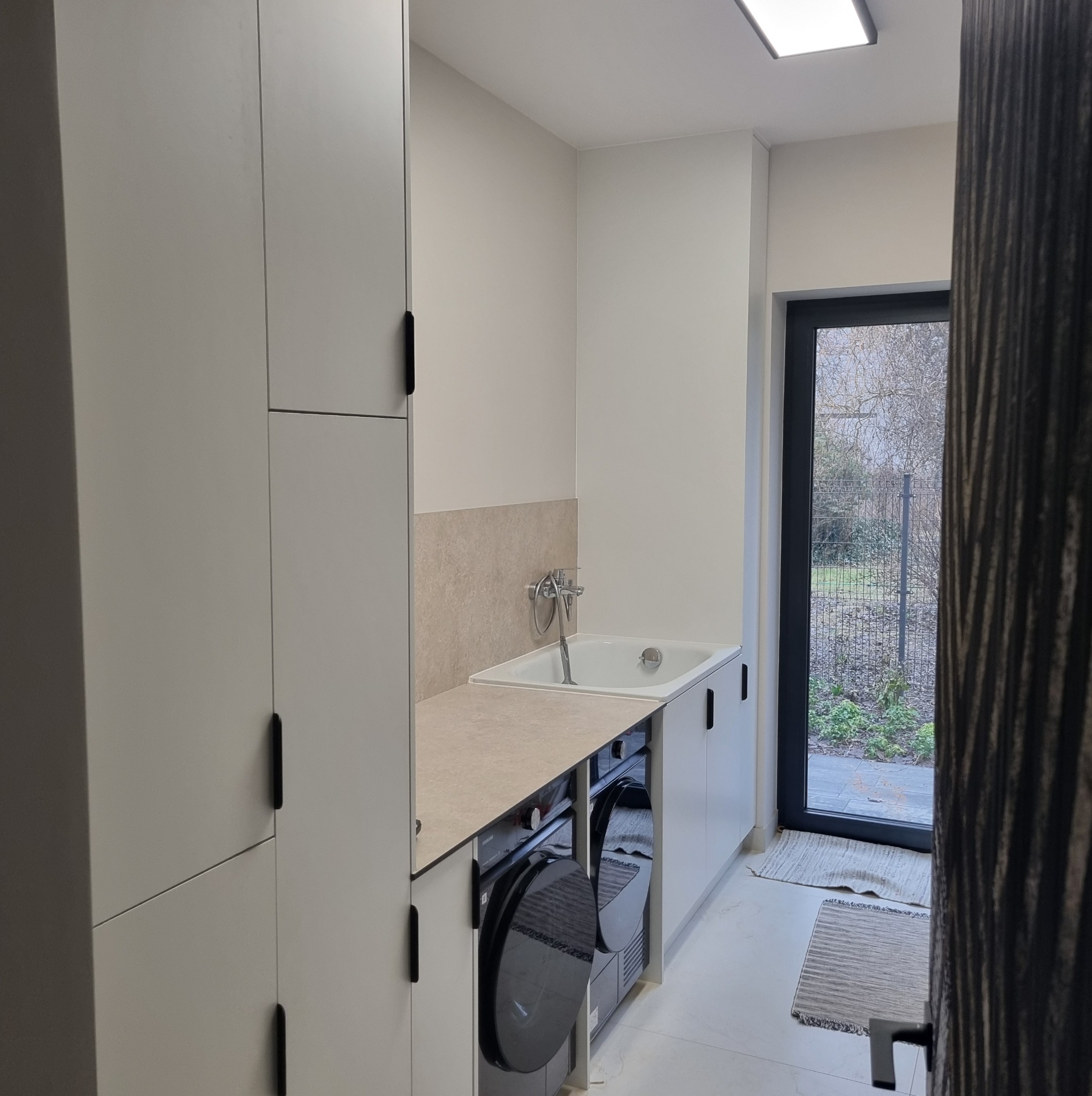 Jasne meble w pralni z pralką i umywalką, minimalistyczne uchwyty, widok na ogród przez drzwi balkonowe, neutralna kolorystyka, czysty design.