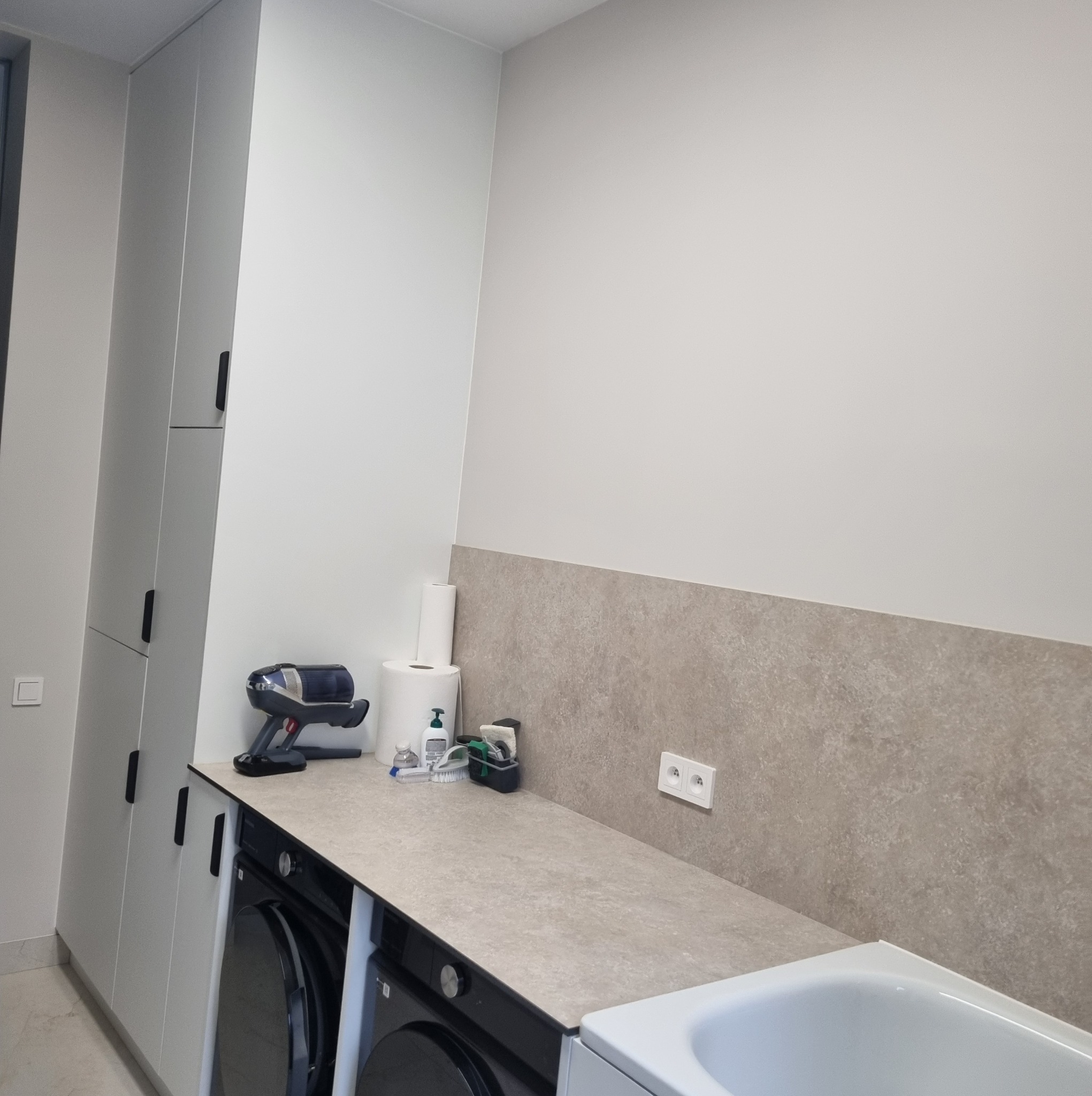 Jasne, wysokie szafy z czarnymi uchwytami w pralni, nad pralką blat z akcesoriami i szarym panelem ściennym. Minimalistyczne wnętrze.