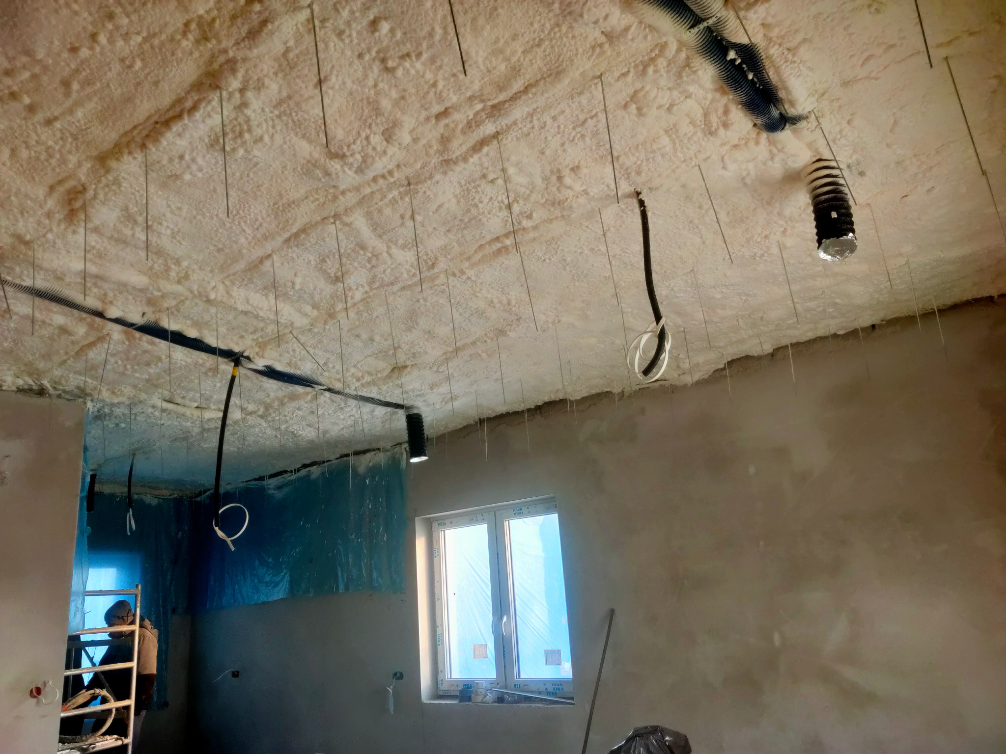 Poddasze w trakcie ocieplania pianką PUR, widoczne przewody wentylacyjne i kable elektryczne zwisające z sufitu, oraz cienkie metalowe pręty stabilizujące pianę.
