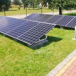 KlimWos - Technology - Dwa rzędy paneli fotowoltaicznych na trawniku w słoneczny dzień, widoczne lampy ogrodowe wzdłuż chodnika z kostki.