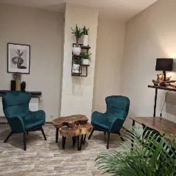 Pokój z dwoma zielonymi fotelami, stolikami kawowymi z plastrów drewna, roślinami doniczkowymi na półce i lampą z drewnianą podstawą, tworzący przytulną przestrzeń do rozmów.