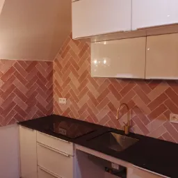 Kuchnia po remoncie: ściana wyłożona ceglastymi płytkami ułożonymi w jodełkę, czarny blat, białe szafki wiszące i stojące, złota bateria kuchenna, gniazdka elektryczne na ścianie.