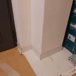 Zamontowane białe listwy przypodłogowe przy ścianie i ościeżnicy, widoczne narzędzia i resztki materiałów budowlanych na podłodze.