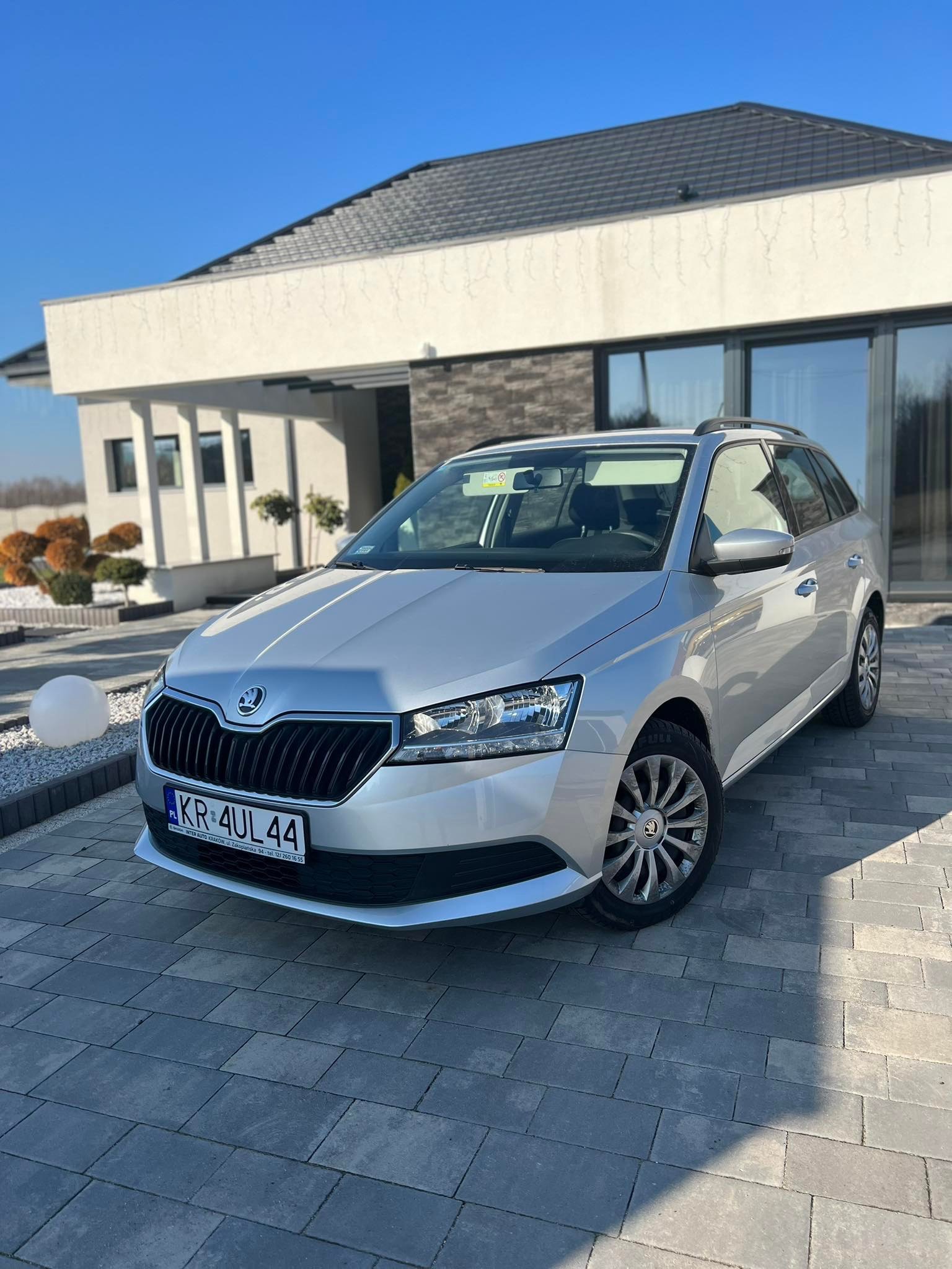 Srebrna Skoda Fabia Combi zaparkowana na brukowanym podjeździe przed nowoczesnym domem z dużymi oknami i płaskim dachem w słoneczny dzień.