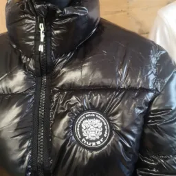 Błyszcząca, czarna kurtka puchowa z naszywką z logo na manekinie, widoczny zamek błyskawiczny z jasnymi detalami.