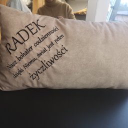 CustoMove - Beżowa poduszka z czarnym napisem 'RADEK Nasz bohater codzienności dzięki Niemu, świat jest pełen życzliwości' leżąca na ciemnym tle, widoczne odbicie osoby w tle.