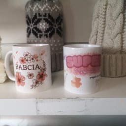CustoMove - Dwa białe kubki z różnymi nadrukami (kwiaty i napis 'Babcia' oraz napis 'Super Przyjaciółka') stoją na białej półce, w tle widać dwie wełniane czapki.
