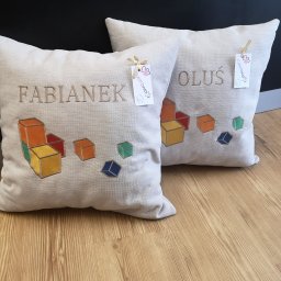 CustoMove - Dwa beżowe poduszki dekoracyjne z haftowanymi imionami (Fabianek i Oluś) oraz motywem kolorowych klocków, leżące na drewnianej podłodze, z metkami producenta przywiązane wstążką.